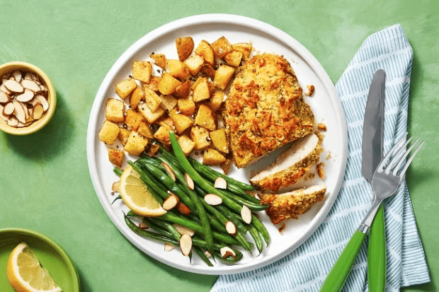 Screenshot 2025-04-07 at 7.05.43 PM Assiette d’un plat HelloFresh avec poulet croustillant, pommes de terre rôties et haricots verts aux amandes, sur fond vert.
