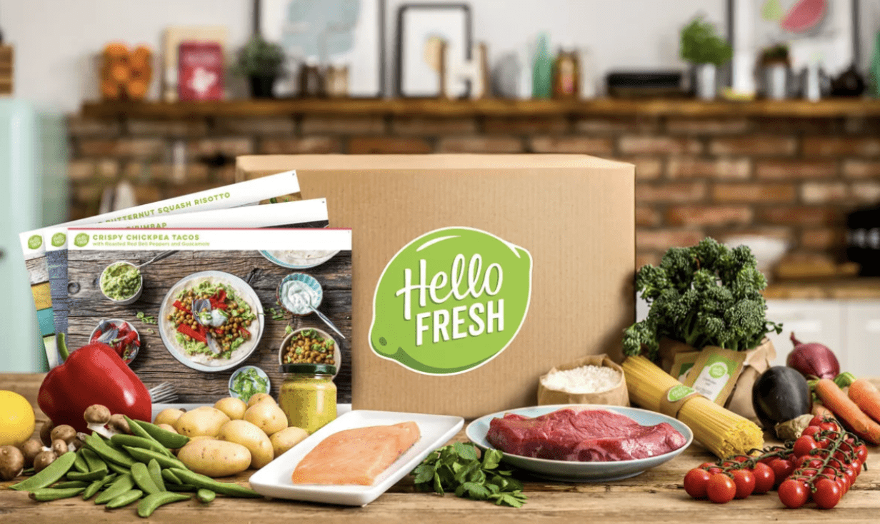Screenshot 2025-04-23 at 1.01.34 PM Box repas HelloFresh ouverte sur un plan de travail, entourée d’ingrédients frais (viande, poisson, légumes, féculents) et de fiches recettes comme « Crispy Chickpea Tacos »