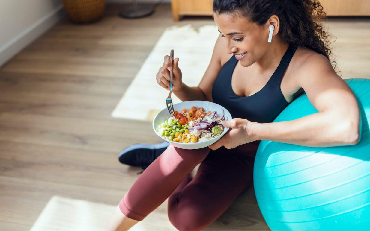 Screenshot 2025-04-27 at 1.51.40 PM Femme en tenue de sport mangeant un repas équilibré après une séance d'entraînement, assise près d'un ballon de gym