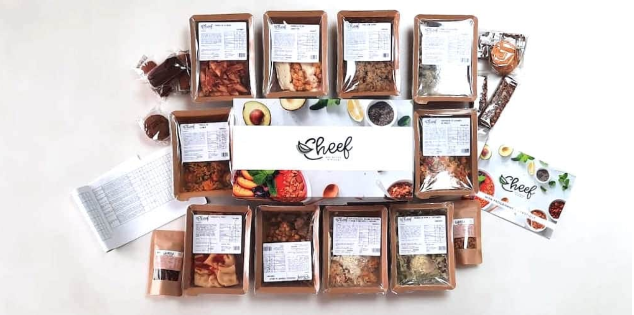Screenshot 2025-06-10 at 6.39.35 PM Box repas Cheef contenant des plats cuisinés emballés individuellement, accompagnés de fiches nutritionnelles, snacks et livret de présentation, disposés autour d’un coffret central au logo Cheef