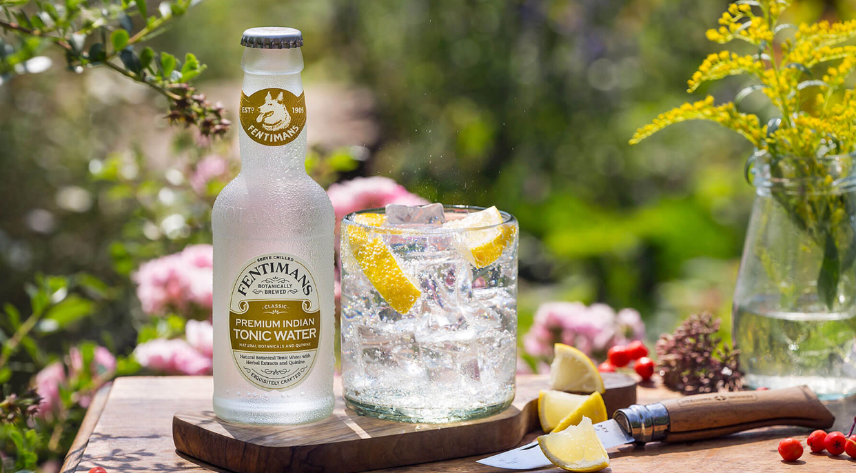 Screenshot 2025-06-13 at 1.19.14 PM Bouteille de tonic Fentimans Premium Indian accompagnée d’un verre rempli de glaçons et de quartiers de citron, posés sur une planche en bois dans un jardin fleuri