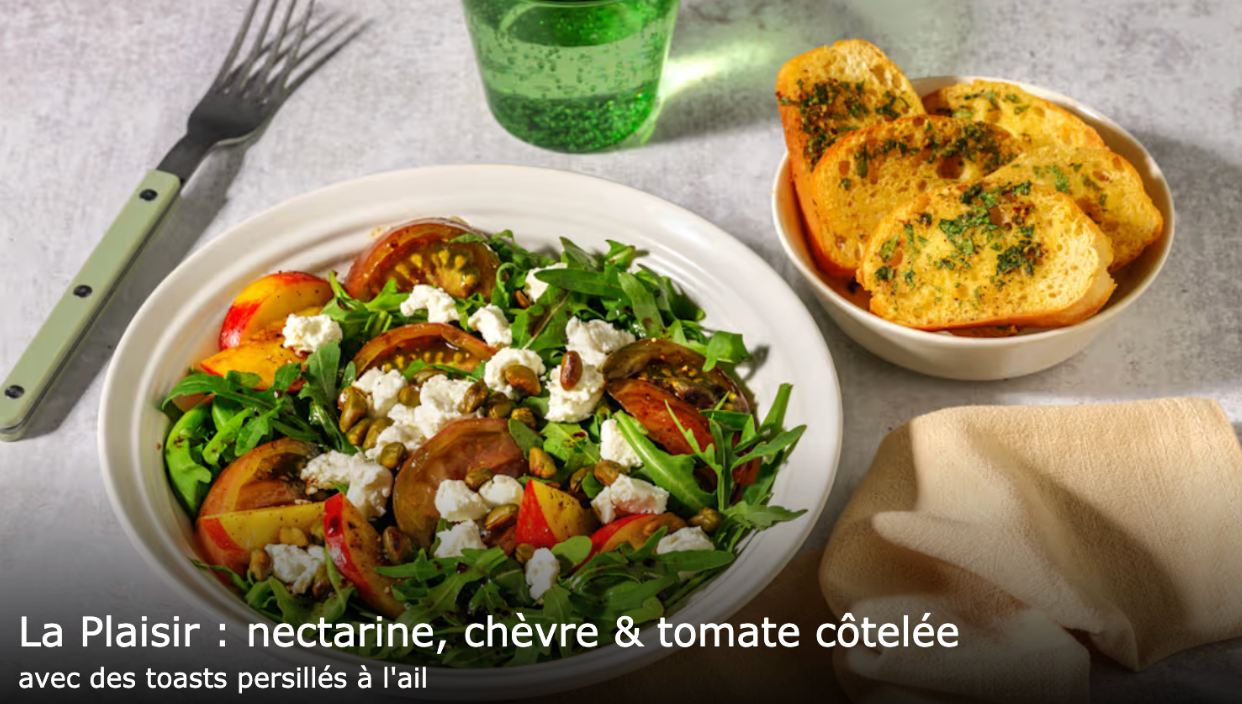 Assiette de salade fraîche composée de roquette, nectarines, tomates côtelées, fromage de chèvre et pistaches, accompagnée de toasts à l’ail et au persil dans un bol, avec une fourchette et un verre d’eau pétillante à l’arrière-plan