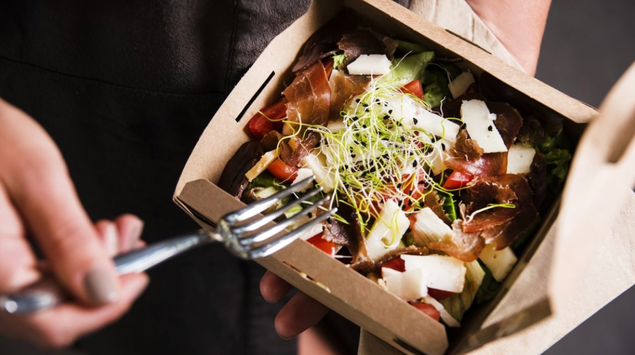 Une main tient une boîte en carton contenant une salade composée de lamelles de viande séchée, de dés de fromage, de tomates, de pousses germées et de feuilles de salade verte. Une fourchette est sur le point de piquer un morceau. Plat minceur typique de Dietbon