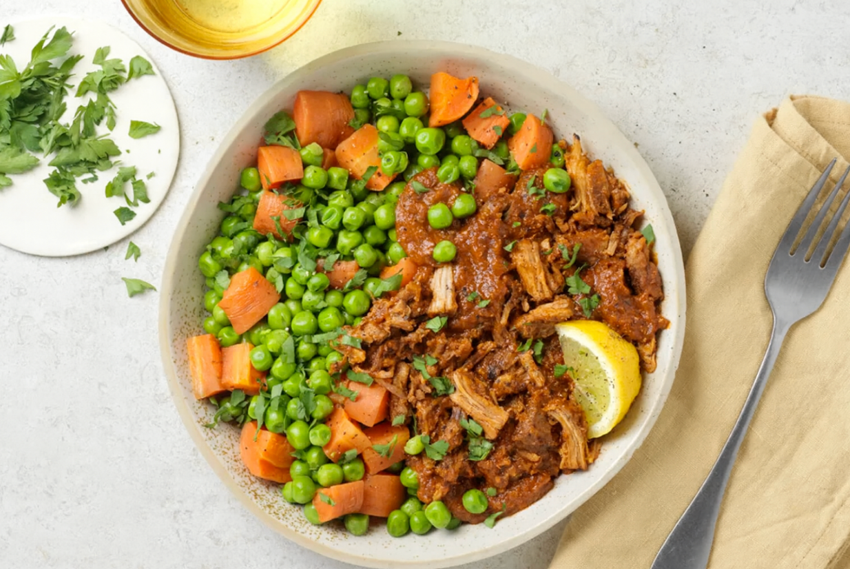 Assiette composée de petits pois, de carottes coupées en morceaux et de viande effilochée en sauce tomate, garnie de coriandre fraîche et d’un quartier de citron. Plat minceur équilibré typique de Seazon, servi avec une fourchette et une serviette beige.