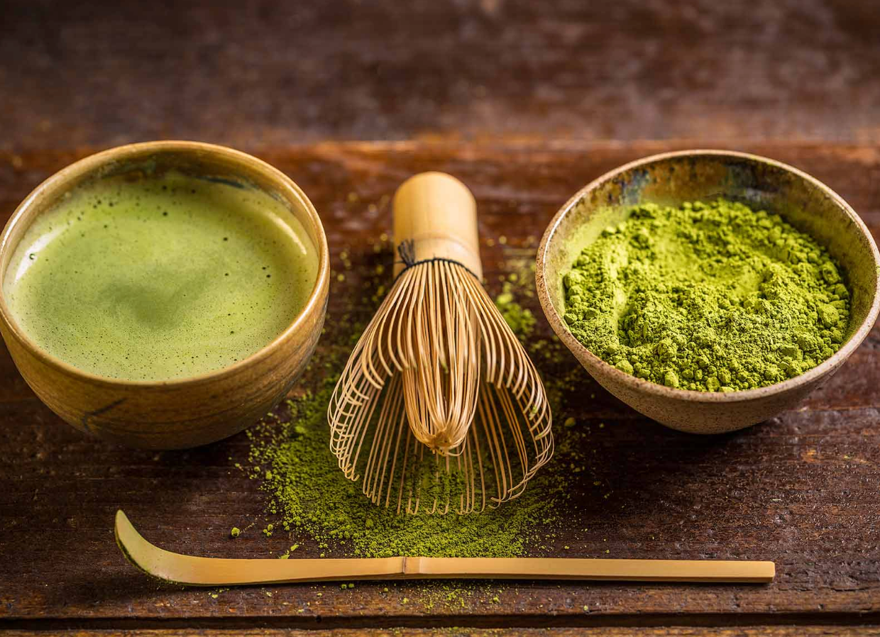 Bol de matcha fouetté, poudre de matcha dans un autre bol, fouet en bambou (chasen) et spatule en bambou (chashaku) posés sur une table en bois.
