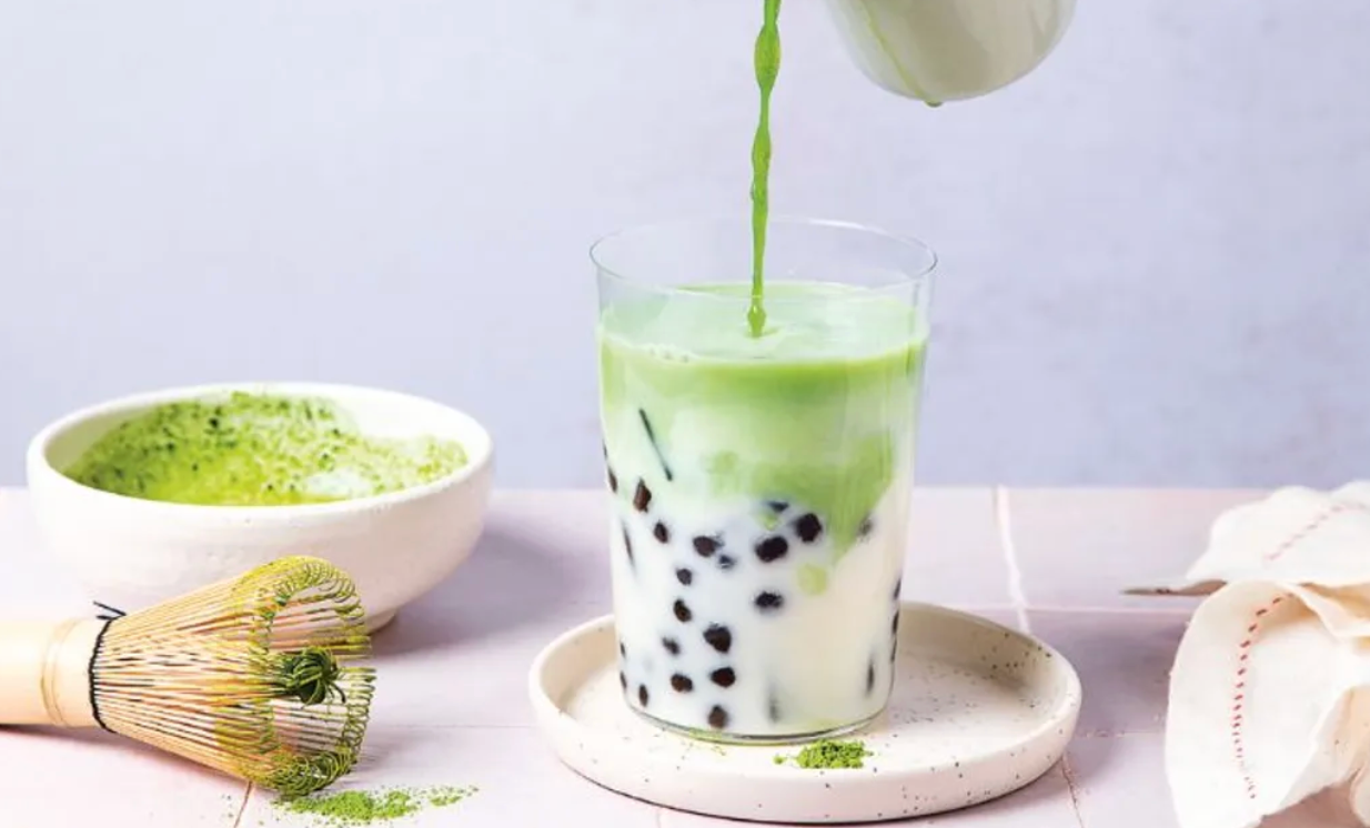 Verre de bubble tea au matcha avec perles de tapioca noires, lait et matcha versé par-dessus, accompagné d’un bol de matcha fouetté et d’un fouet en bambou (chasen) sur une table claire
