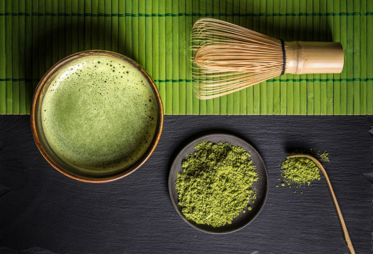 Vue de dessus d’un bol de matcha préparé, posé sur un napperon vert, accompagné d’un fouet en bambou (chasen), d’une assiette de poudre de matcha et d’une spatule en bambou (chashaku) sur une surface noire.