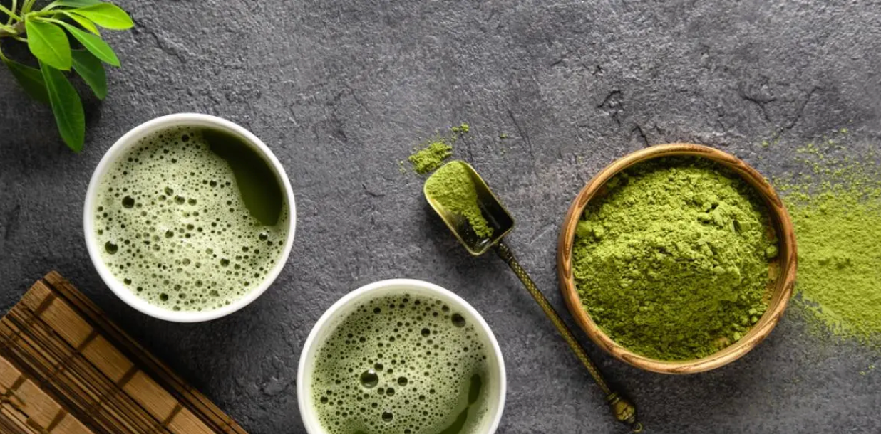 Deux tasses de thé matcha mousseux vues de dessus, accompagnées d’un bol en bois rempli de poudre de matcha, d’une cuillère en métal contenant du matcha et de feuilles vertes décoratives sur une surface en pierre grise