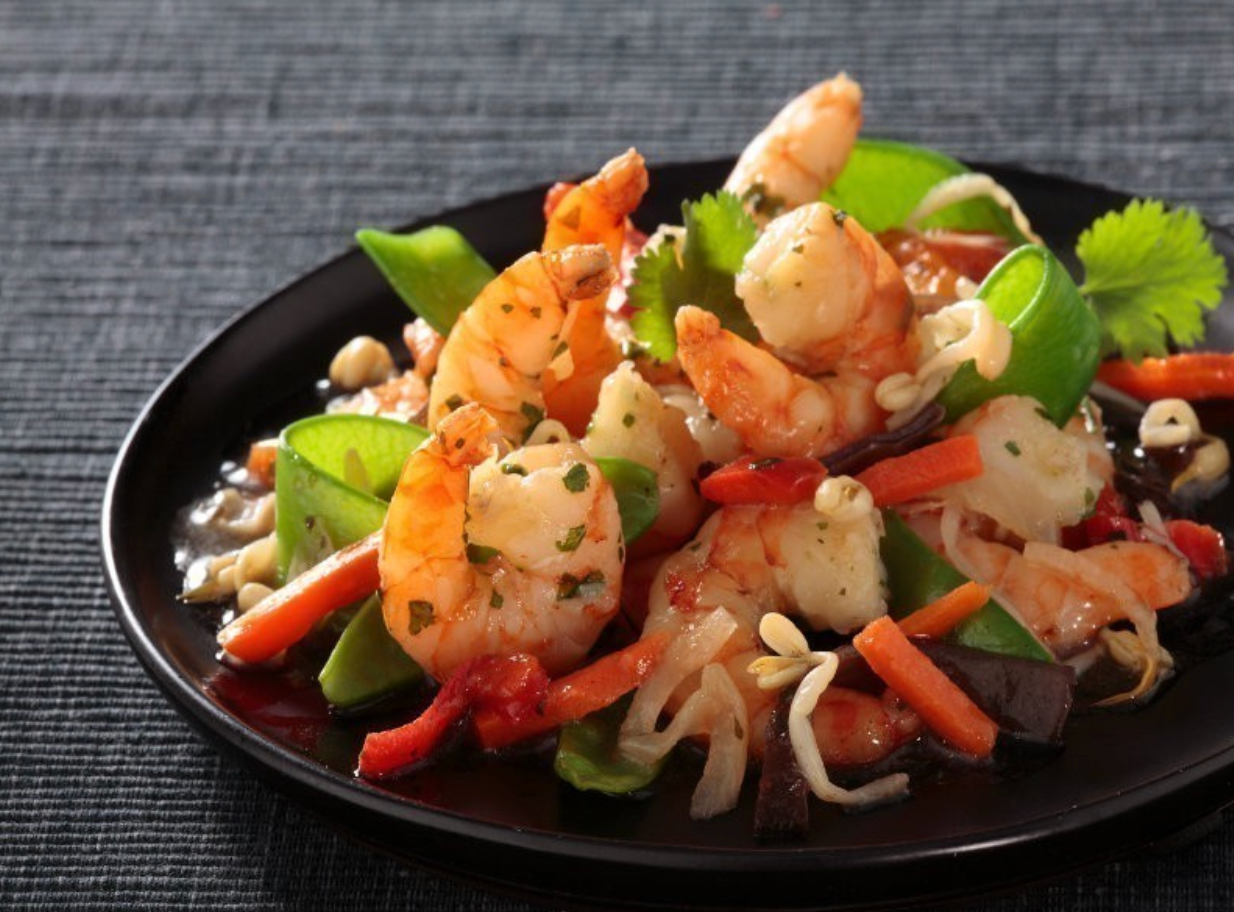 Screenshot 2025-07-10 at 4.02.54 PM Plat cuisiné aux crevettes, légumes croquants et sauce asiatique, servi avec coriandre et citron vert sur une assiette noire.