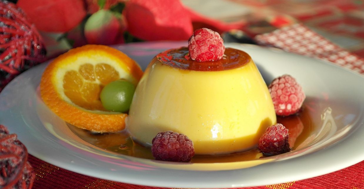 Flan au caramel décoré de framboises, accompagné d’une tranche d’orange et d’un raisin vert, servi sur une assiette blanche