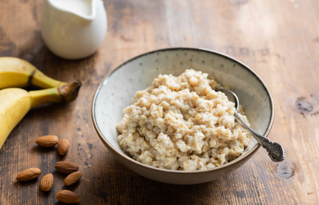 Bol de flocons d’avoine cuits, accompagné de bananes, d’amandes et d’un pichet de lait, sur une table en bois