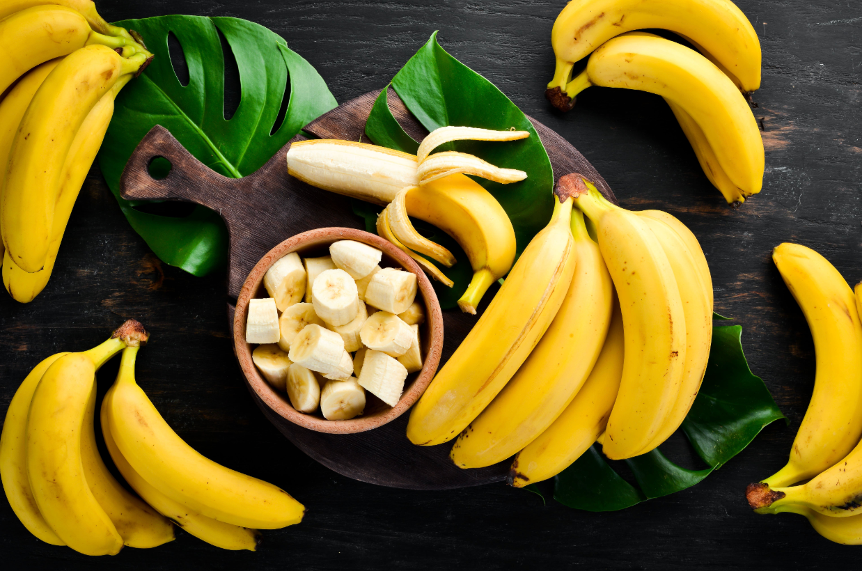 Bananes fraîches entières et coupées en rondelles dans un bol en bois, disposées sur une planche avec des feuilles vertes, sur fond sombre.