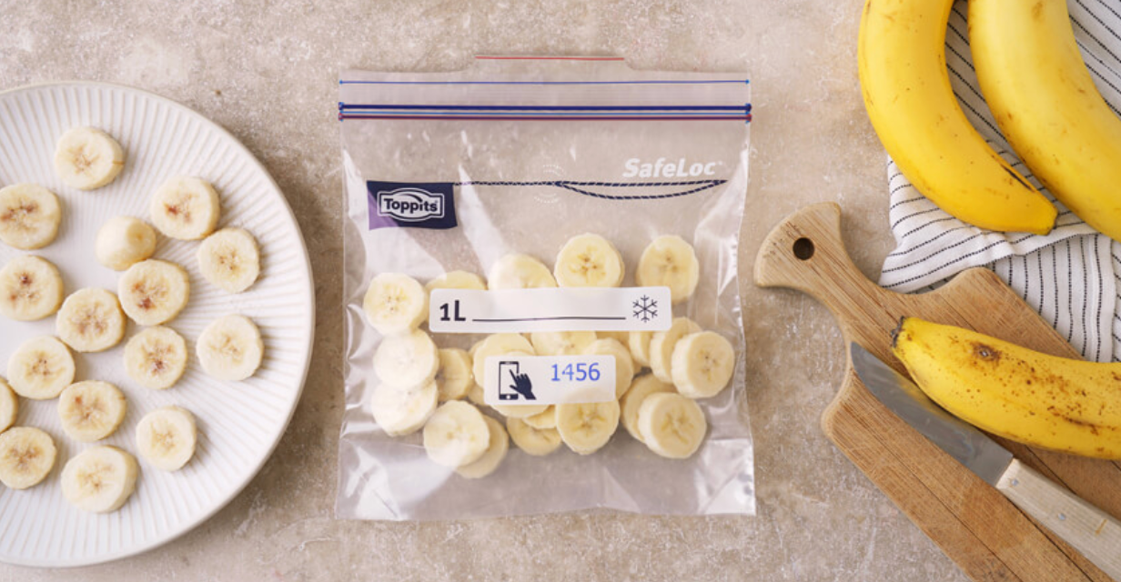 Rondelles de banane dans un sachet de congélation transparent, avec une assiette de bananes tranchées, une planche à découper et des bananes entières