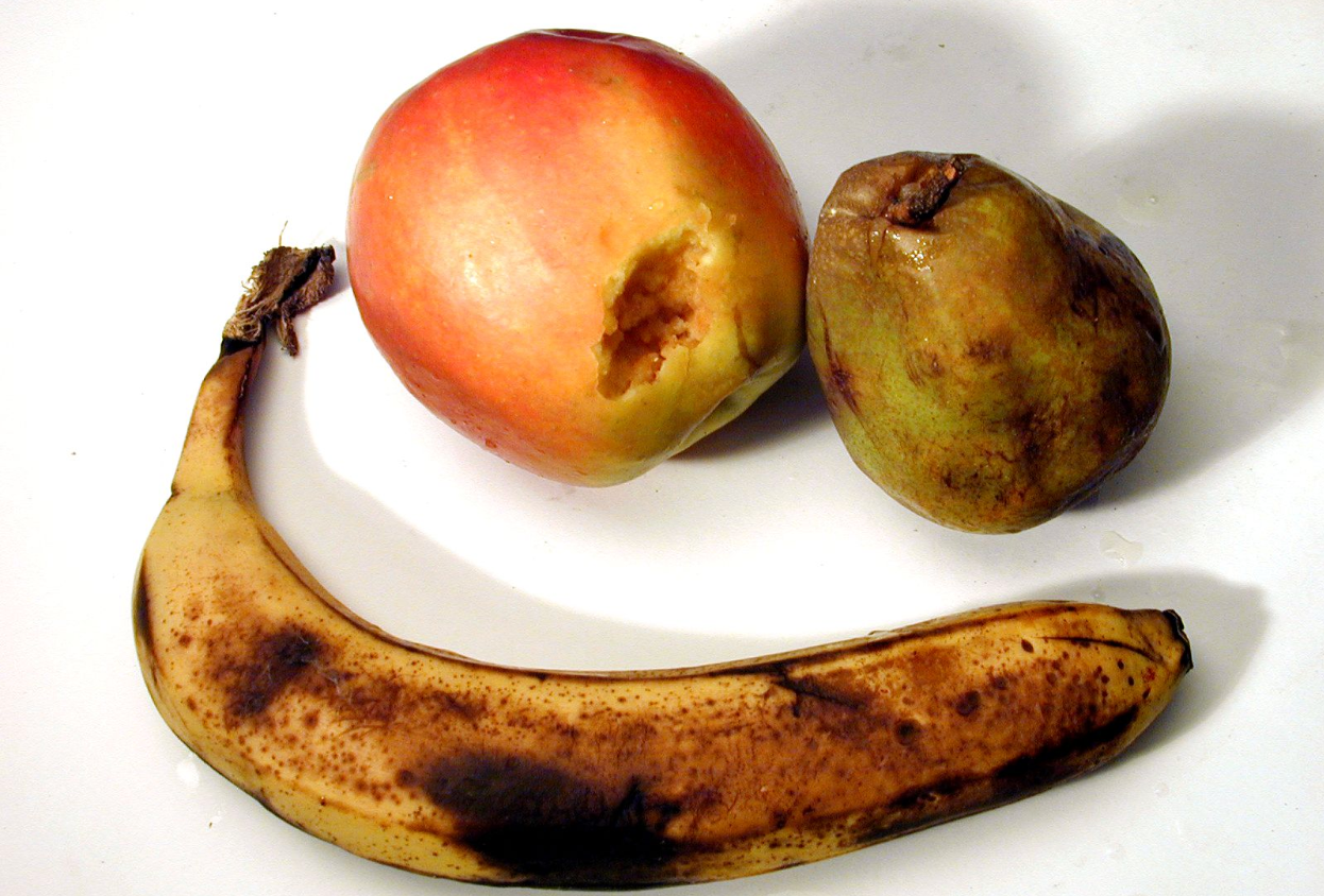 Fruits abîmés sur une table : une banane très mûre avec des taches brunes, une pomme avec une partie entamée et une poire abîmée