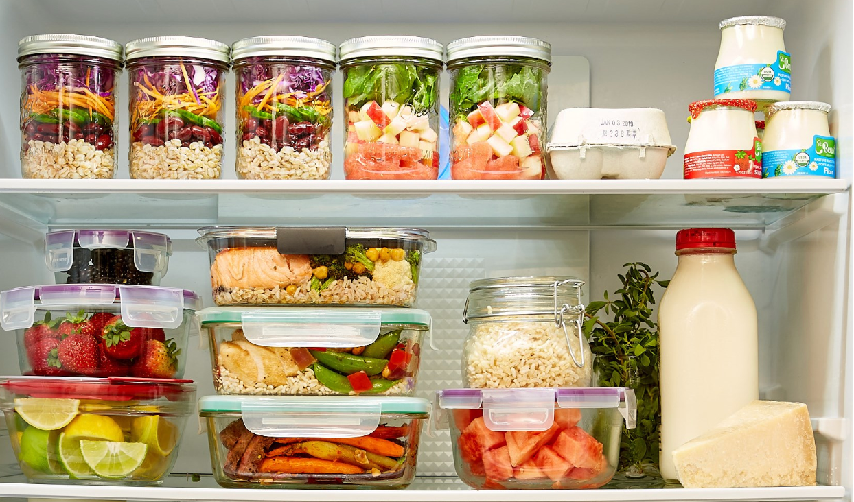 Réfrigérateur bien organisé contenant des bocaux de salades colorées et de céréales, des plats préparés dans des boîtes hermétiques (saumon, légumes, riz), des fruits frais (fraises, citrons, pastèque), des yaourts, du lait, du fromage et des herbes aromatiques