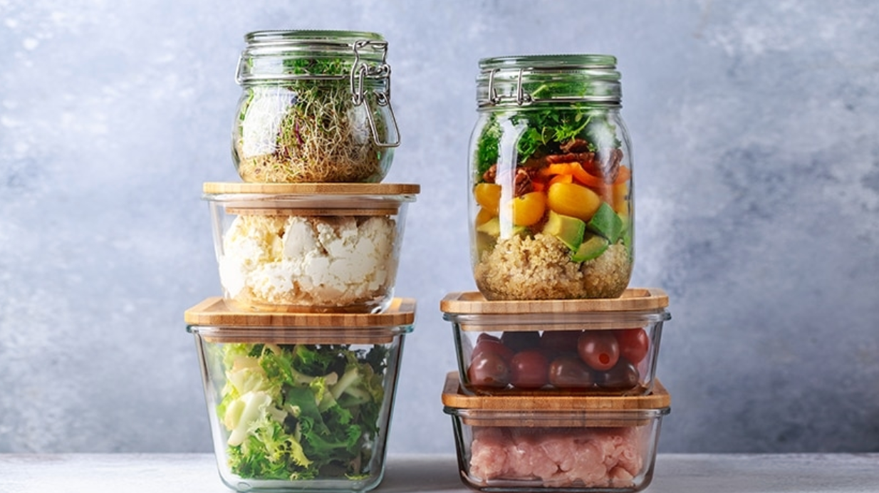 Ingrédients de box repas conservés dans des bocaux et boîtes hermétiques en verre avec couvercles en bambou, contenant légumes, quinoa, fromage, tomates et germes, posés en pile sur une table